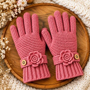 May include: A pair of rose pink crochet gloves featuring a ribbed cuff, a decorative strap with a central crocheted flower, and a small circular wooden button with a floral cutout pattern on each wrist, showcasing a textured knit construction.
