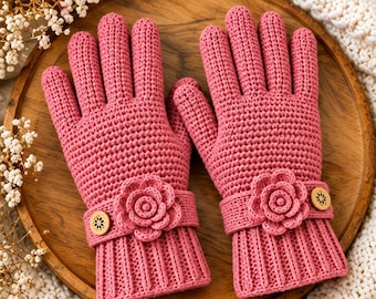 May include: A pair of rose pink crochet gloves featuring a ribbed cuff, a decorative strap with a central crocheted flower, and a small circular wooden button with a floral cutout pattern on each wrist, showcasing a textured knit construction.