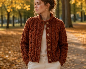 May include: A rust-colored cable knit cardigan featuring a high collar, long sleeves, and a front button closure with eight light-colored circular buttons. The garment is worn over a plain white shirt and light-colored trousers.