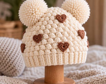 May include: A cream-colored crocheted beanie featuring a textured bobble stitch pattern, two large matching pom-poms on top, and several small, dark brown crocheted heart-shaped appliques scattered across the surface of the hat.