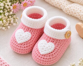 May include: A pair of handmade crochet baby booties in a soft pink color. Each bootie features a white crocheted heart appliqué on the toe, a white ribbed cuff, and a single light brown wooden button attached to the side of the ankle collar.