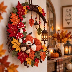 May include: A crochet wreath featuring a sleeping orange and white fox, red and white mushrooms, brown acorns, and vibrant autumn leaves in red, orange, and green. The wreath includes hanging wooden beads and a small lantern with a warm light.