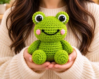 May include: A small, bright green crocheted frog plush toy. The frog features large, round black eyes with white borders, a stitched black smile, and small pink circular patches on its cheeks. The toy has a rounded head and body with stubby limbs.
