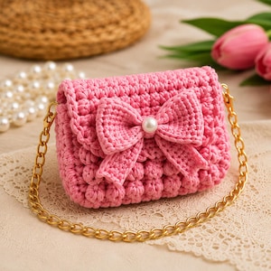 Può includere: Una piccola borsa a mano all'uncinetto di colore rosa con un motivo a punti a rilievo e un grande fiocco all'uncinetto in tinta sulla patta anteriore. Il centro del fiocco è decorato con una perla bianca. Include una tracolla a catena dorata.