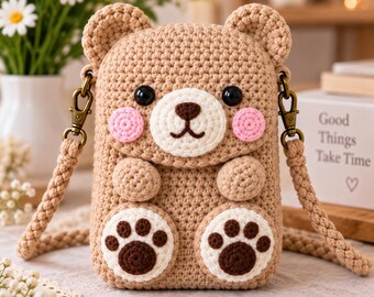 May include: A light brown crochet bear bag with a cream-colored muzzle, dark brown nose, and pink cheeks. The bag features two small ears, two arms, and two feet with dark brown paw pads. It has a braided strap attached with bronze-colored clips.