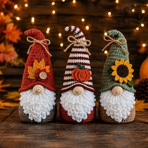 May include: Three handmade crochet gnomes with white beards and round noses. The left gnome wears a red hat with a maple leaf and button. The centre gnome has a striped hat with a pumpkin. The right gnome features a green hat with a sunflower.