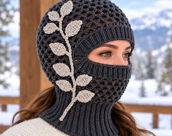 May include: A dark gray crochet balaclava featuring an open mesh stitch pattern and a ribbed neck opening. The headpiece is decorated with a light beige crocheted branch applique with multiple leaves extending along the side of the face.