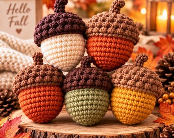 May include: A collection of five crocheted acorns featuring textured brown caps and bodies in cream, orange, and light green colors. Each acorn has a small stem on top, arranged in a cluster to display the varied yarn colors and stitch patterns.