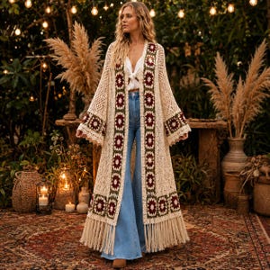 May include: A long, cream-colored crochet duster cardigan featuring a repeating granny square pattern in dark red and olive green along the front panels and sleeves. The garment has wide, open sleeves, a floor-length hem, and long fringe trim.