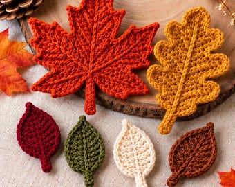 May include: A collection of six crocheted leaves in various shapes and colors, including a large orange maple leaf, a golden yellow oak leaf, and four smaller oval leaves in deep red, olive green, cream, and brown, all featuring visible yarn texture.