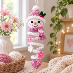 May include: A crocheted snowman ornament with a pink hat, a matching scarf featuring a white snowflake, and a spiral body. The snowman is decorated with small pink and white flowers and ends with a pink heart charm labeled with the word JOY.