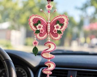 May include: A crocheted butterfly car ornament featuring pink and cream yarn, accented with small white pearls, green leaves, and pink flowers. A spiral tail hangs below the butterfly, ending in a faceted pink crystal drop pendant.