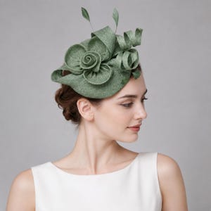 May include: A sage green fascinator hat featuring a textured fabric base, a central rose-shaped floral accent, and structured ribbon loops. Two thin, upright feather-like stems extend from the top of the headpiece, which is worn at an angle.