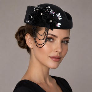 May include: A black pillbox hat featuring a cluster of black fabric flowers adorned with clear crystals and iridescent accents. The hat includes a decorative arrangement of delicate black beaded chains that drape down the side of the headpiece.