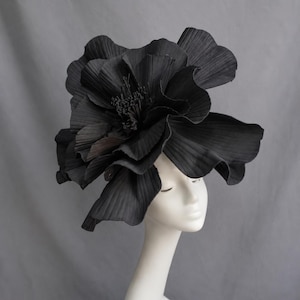 Puede incluir: Un tocado floral negro de gran tamaño con pétalos plisados en capas y un centro texturizado. El accesorio se exhibe sobre una cabeza de maniquí blanca y lisa, destacando los bordes ondulados y la estructura detallada del diseño floral oscuro.