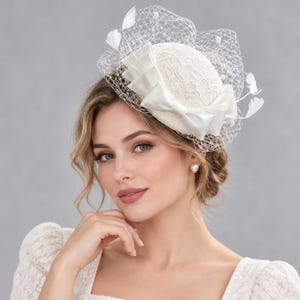 May include: A cream-colored fascinator hat featuring a lace-covered dome, a satin ribbon bow, and a white birdcage veil. The headpiece is adorned with small white feathers and pearl accents, creating a structured and layered appearance.
