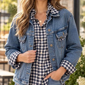 May include: A blue denim jacket with metal buttons and two chest pockets, worn over a navy and white gingham button-down shirt with a ruffled collar. The jacket features long sleeves with folded cuffs revealing the gingham pattern underneath.
