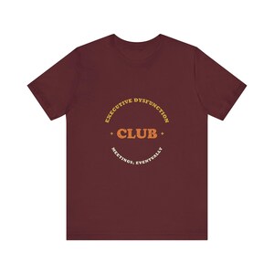 May include: A maroon short-sleeved t-shirt featuring a circular graphic in yellow, orange, and white. The text reads "EXECUTIVE DYSFUNCTION CLUB" with "MEETINGS: EVENTUALLY" printed below. The design includes small star accents on the sides.