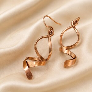 May include: A pair of copper-toned dangle earrings featuring a twisted, ribbon-like spiral design. Each earring is suspended from a simple curved hook, showcasing a metallic finish with subtle variations in color and a smooth, flowing shape.