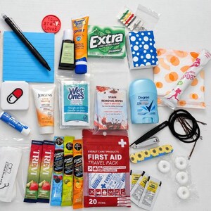 May include: A collection of travel-sized personal care items including a blue notepad, black pen, sunscreen, Extra gum, stain wipes, Degree deodorant, Trevi drink mixes, energy sticks, a first aid kit, hair ties, bobby pins, and mints.