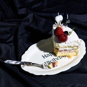 Könnte beinhalten: Ein Stück Schichtkuchen mit weißer Glasur, garniert mit einer Erdbeere, einer Kirsche und zwei herzförmigen Kerzen, eine weiß und eine schwarz mit dem Text Happy Birthday. Der Kuchen liegt auf einem ovalen weißen Teller mit dem Text Happy Birthday.