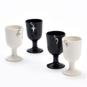 Könnte beinhalten: Vier keramische Stielbecher in Schwarz und Weiß, jeweils mit einem silberfarbenen Metallanhänger am Rand. Die Anhänger umfassen zwei sternförmige Designs, eine Zuckerstange und eine Schneeflocke, die an kleinen silbernen Ringen befestigt sind.