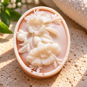 May include: An oval-shaped cameo brooch featuring a raised, white, carved floral relief design set against a smooth, light peach-colored background. The intricate carving depicts a cluster of blooming flowers and leaves with fine, detailed petals.