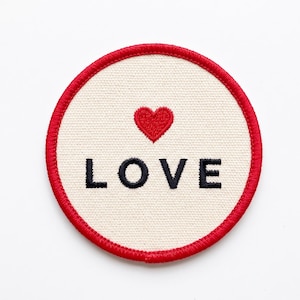 May include: A circular embroidered patch with a cream-colored fabric base and a vibrant red stitched border. Centered above the black embroidered text that reads LOVE is a small, solid red heart icon, creating a clean and simple design.