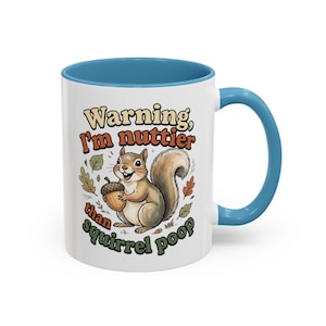 May include: A white ceramic mug with a blue interior and handle, featuring a cartoon squirrel holding an acorn. The text reads: Warning, I'm nuttier than squirrel poop. The design includes small colorful autumn leaves surrounding the squirrel.