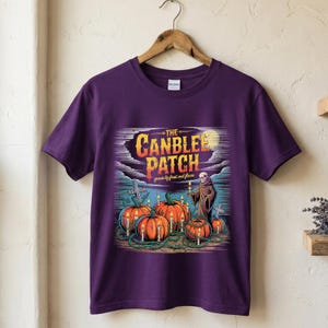 May include: A purple short-sleeve t-shirt featuring a graphic print of orange pumpkins, lit candles, and a skeleton figure. The text reads THE CANBLEE PATCH grown by frost and flame. The design includes a graveyard scene with crosses and a full moon.