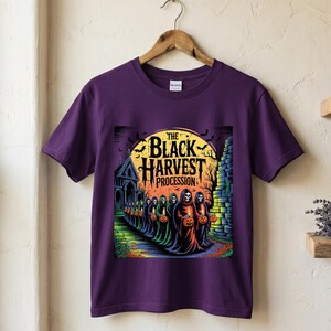 May include: A purple short-sleeve t-shirt featuring a graphic print of a procession of hooded skeletons carrying glowing orange jack-o'-lanterns in front of a large yellow moon, a church, and bats, with the text THE BLACK HARVEST PROCESSION.