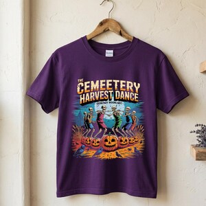 May include: A purple short-sleeve t-shirt featuring a graphic print of skeletons dancing in a circle around pumpkins. The text reads THE CEMEETERY HARVEST DANCE and BONES BY MOONLIGHT. The design includes a full moon, crows, and tall grass.
