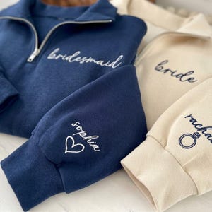 May include: A navy blue quarter-zip sweatshirt with white embroidered text reading bridesmaid and the name sophia with a heart icon on the sleeve. Beside it, a beige quarter-zip sweatshirt features dark text reading bride and rachel with a ring icon.