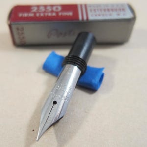 May include: A silver fountain pen nib with the text "2550" and a black section. A blue cloth rests beneath the nib. The pen is in front of a rectangular metal box with the text "2550 Firm Extra Fine".