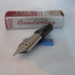 May include: A silver fountain pen nib with a black section, resting on a blue support. The nib is in focus, with the words "Broad writing" visible on the packaging in the background. The packaging is red and white.