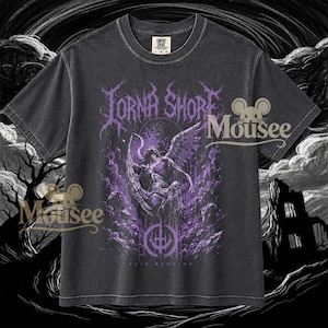 Puede incluir: Una camiseta gris carbón de manga corta con un estampado gráfico morado de una figura alada y el texto Lorna Shore y Pain Remains. La camiseta tiene cuello redondo y una etiqueta de la marca Comfort Colors visible en el interior del cuello.