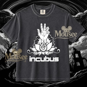 Puede incluir: Una camiseta gris carbón de manga corta con un gráfico blanco de una mano emergiendo de una flor de loto. La palabra "incubus" está impresa en blanco debajo del gráfico. La camiseta tiene cuello redondo y una etiqueta Comfort Colors visible.