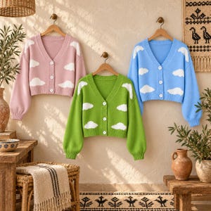 May include: Three V-neck cardigans hanging on wooden hangers, each featuring a white cloud pattern. One cardigan is light pink, one is bright green, and one is light blue. Each sweater has long sleeves, ribbed cuffs, and a front button closure.