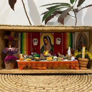 May include: A miniature wooden box diorama featuring a Day of the Dead altar display. Inside, a table with an orange lace cloth holds tiny food items, skulls, and bottles. A central image of Our Lady of Guadalupe is flanked by two white skulls.