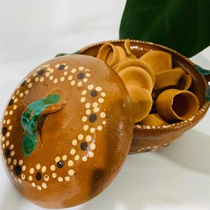 Puede incluir: Una olla de barro marrón con tapa a juego, con un asa verde y patrones florales pintados en blanco y marrón oscuro. La olla está llena de varios cuencos pequeños de barro marrón claro sin esmaltar, cada uno con un borde circular.