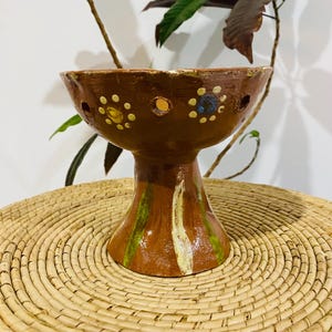 Puede incluir: Un cuenco de cerámica marrón sobre pedestal con un borde ancho y acampanado que presenta una serie de pequeños recortes circulares. El exterior está decorado con motivos florales pintados a mano en amarillo y azul y pinceladas verticales verdes y blancas.