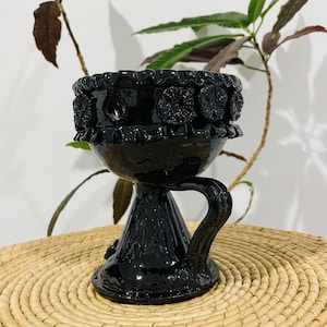 Puede incluir: Un cáliz de cerámica negra brillante con un borde ancho y ondulado, decorado con una serie de motivos florales circulares en relieve. El recipiente tiene una base de pedestal robusta y acampanada, y un asa curva en el lateral.