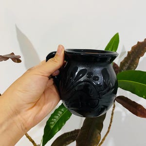Puede incluir: Una taza de cerámica negra de forma redondeada con un acabado brillante y un patrón floral en relieve en su superficie exterior. La taza cuenta con un asa curva en el lateral y un borde circular ancho, presentando un diseño oscuro y monocromático.