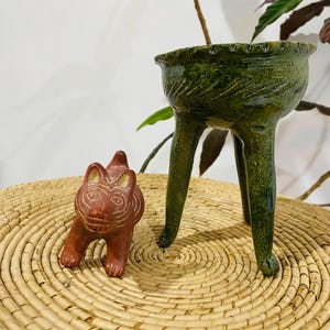 Puede incluir: Una pequeña figura de animal de cerámica color terracota con detalles de líneas incisas y un recipiente trípode de cerámica esmaltada verde con un borde texturizado y tres patas delgadas. Ambos objetos están sobre una superficie tejida.