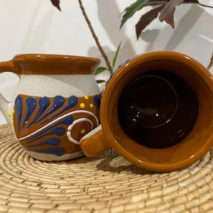 Puede incluir: Dos tazas de cerámica de forma redondeada con borde e interior de color marrón brillante. Una taza muestra un exterior de color crema con motivos florales azules pintados a mano y pequeños puntos amarillos. La otra taza muestra su interior.