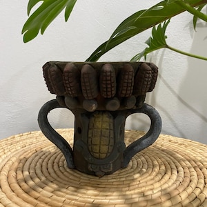 May include: A dark brown ceramic vessel featuring a decorative rim adorned with a series of small, sculpted corn cobs. The body of the pot displays a central corn motif with a gold-toned finish, flanked by two curved handles on either side.