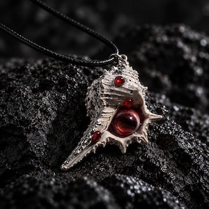 May include: A pendant featuring a textured, off-white seashell shape adorned with small, round, deep red cabochons and a larger, central red eye-like sphere, suspended from a thin black cord necklace.