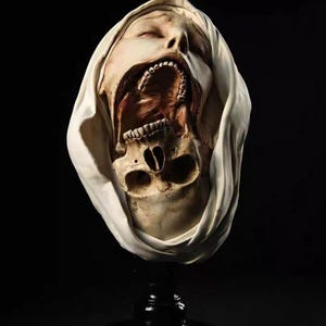May include: A sculpture of a human head draped in a white cloth, featuring a partially skeletal face with an open mouth revealing teeth and a jawbone structure. The skin tone is pale, and the eyes are closed, set against a dark background.