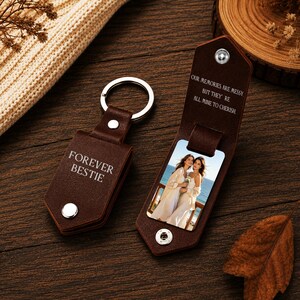 May include: Two dark brown leather keychains with silver metal hardware. One is closed and engraved with the text FOREVER BESTIE. The other is open, revealing a photograph of two people and the engraved text OUR MEMORIES ARE MESSY. BUT THEY'RE ALL MINE TO CHERISH.