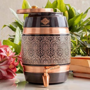 May include: A barrel-shaped copper water dispenser featuring a dark brown finish with an intricate, repeating floral pattern etched into the center. It includes a matching copper lid, a functional spigot, and the text Indian Art Villa A World of Artness.